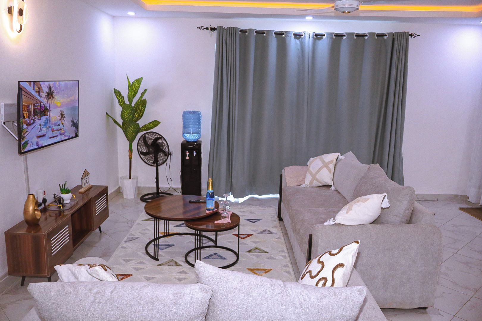 A living room with a sofa, a coffee table, a TV mounted on the wall, a water dispenser, and a fan.