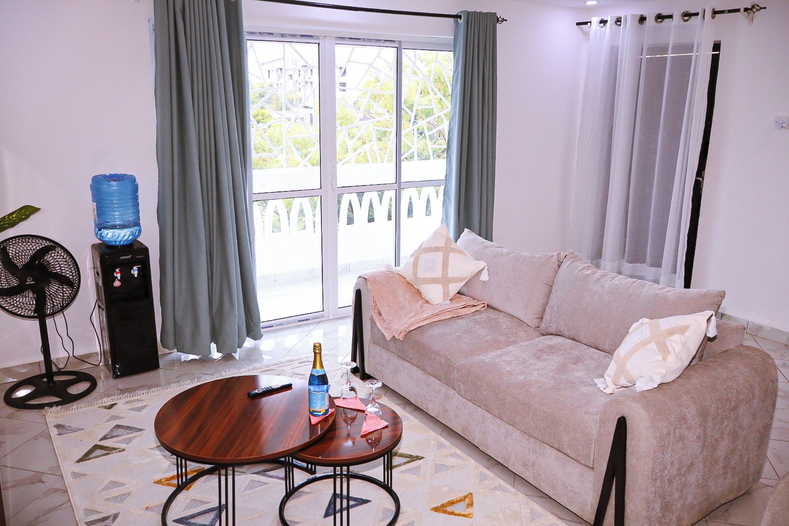A living room with a sofa, a coffee table, a water dispenser, a fan, and a view through a glass door.