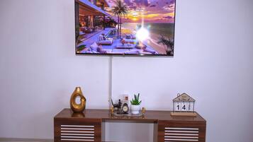 A flat-screen TV mounted on a wall displaying a sunset view. Below the TV, there is a wooden desk with a decorative vase, a small potted plant, a clock, and a calendar.