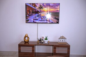 A flat-screen TV mounted on a wall displaying a sunset view. Below the TV, there is a wooden desk with a decorative vase, a small potted plant, a clock, and a calendar. - 2 BEDROOM APARTMENT IN DIANI, UPTO 6 GUESTS C, 5 MINS DRIVE TO THE BEACH (Diani Beach)