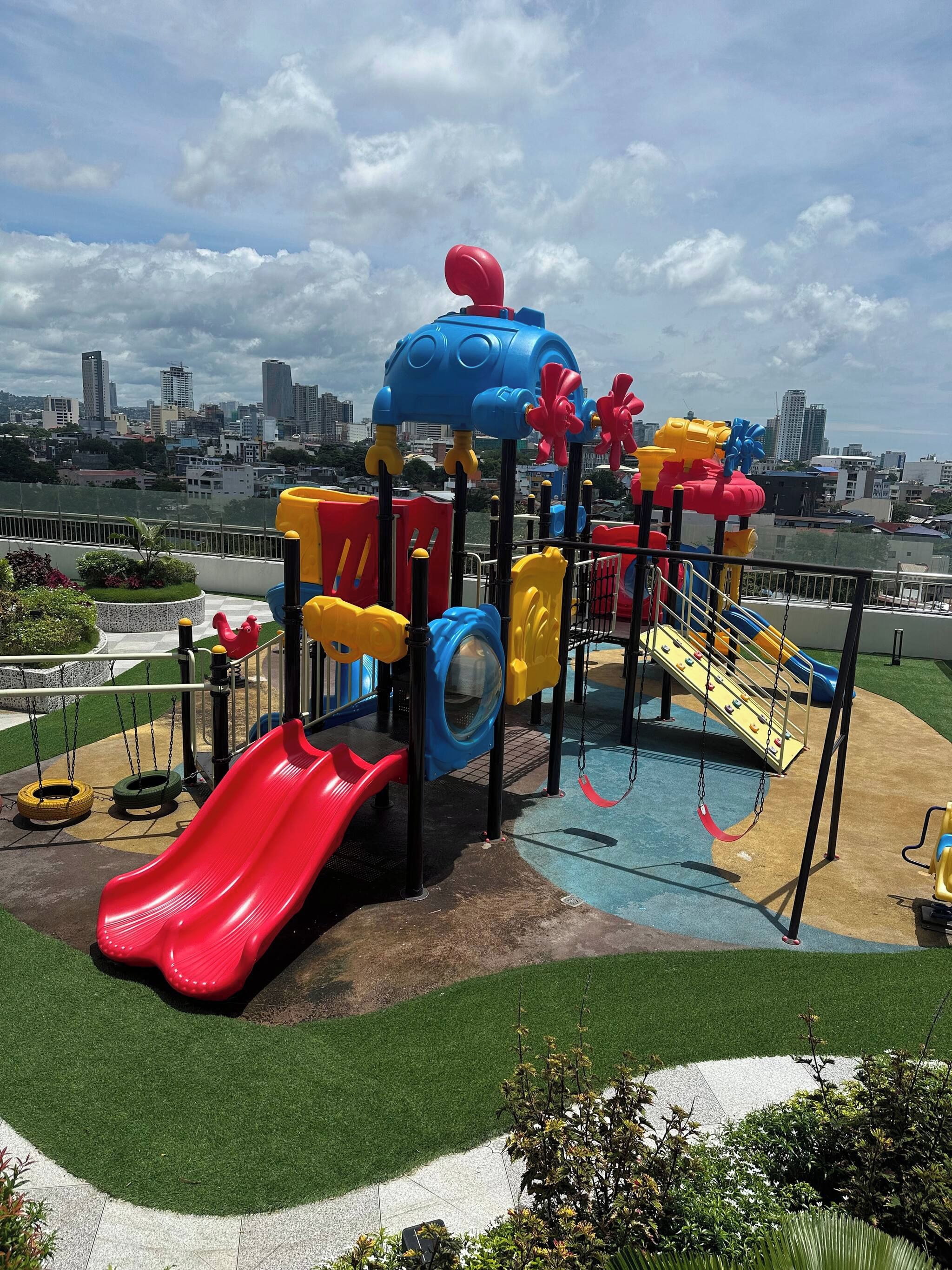 A playground with slides and swings, surrounded by green grass and a cityscape in the background.