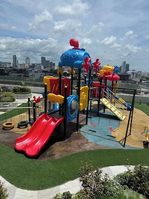 Un parque infantil con toboganes y columpios, rodeado de césped y un paisaje urbano al fondo.