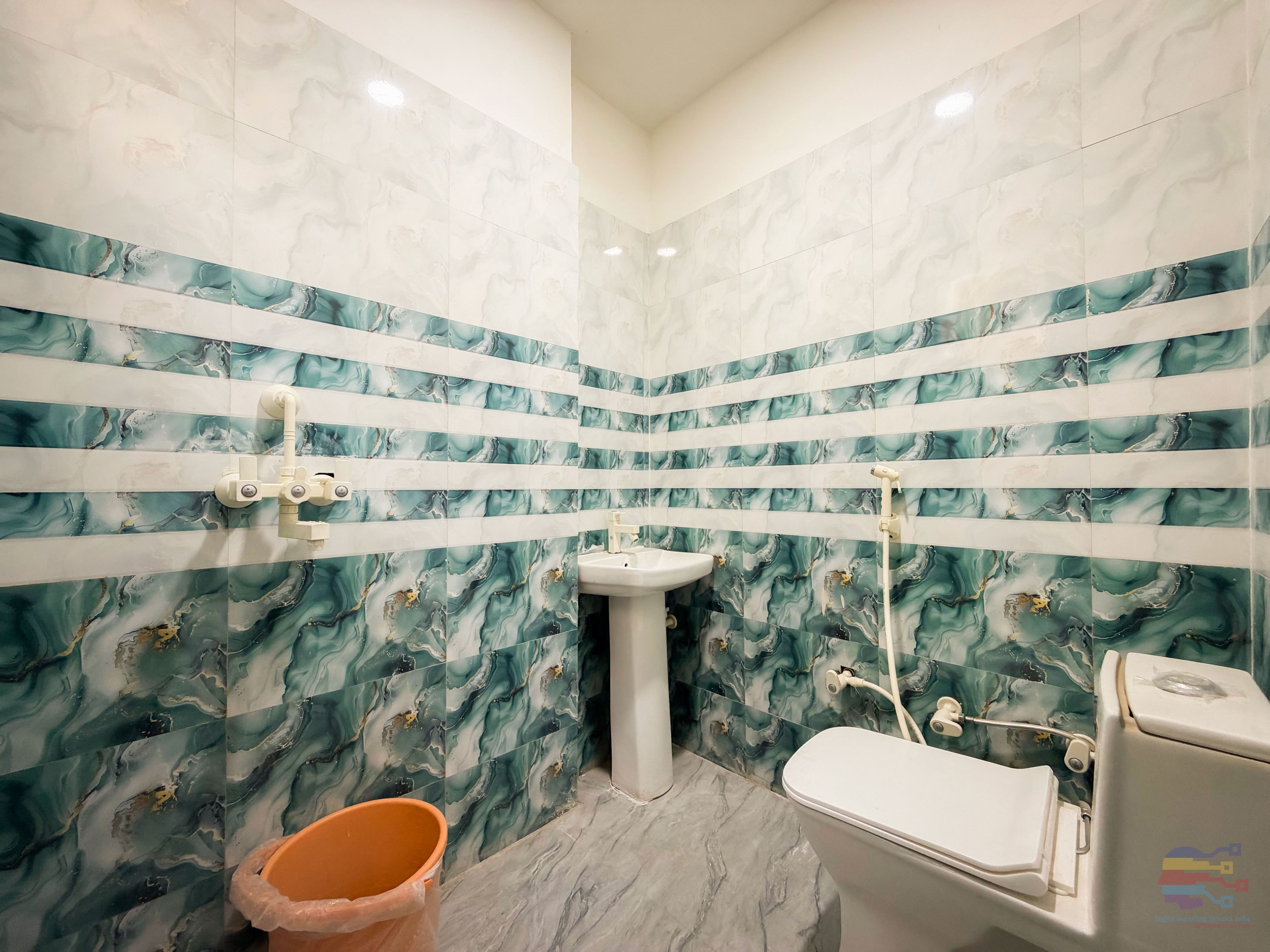 A bathroom with a marble wall design, a pedestal sink, a toilet, and a trash bin.