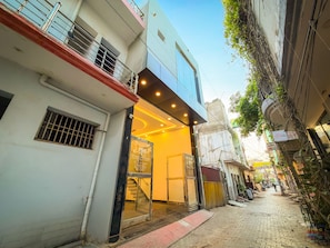A narrow street with buildings on either side, a visible entrance to a property with a glass door, and a metal staircase leading to a balcony. - Shri Kishori Ji Dhaam Homestay (Mathura)