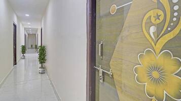 A corridor with a decorative door on the right, potted plants along the left wall, and a ceiling with recessed lighting.