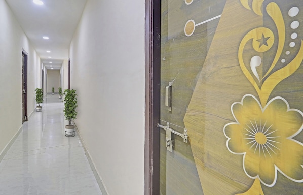 A corridor with a decorative door on the right, potted plants along the left wall, and a ceiling with recessed lighting.