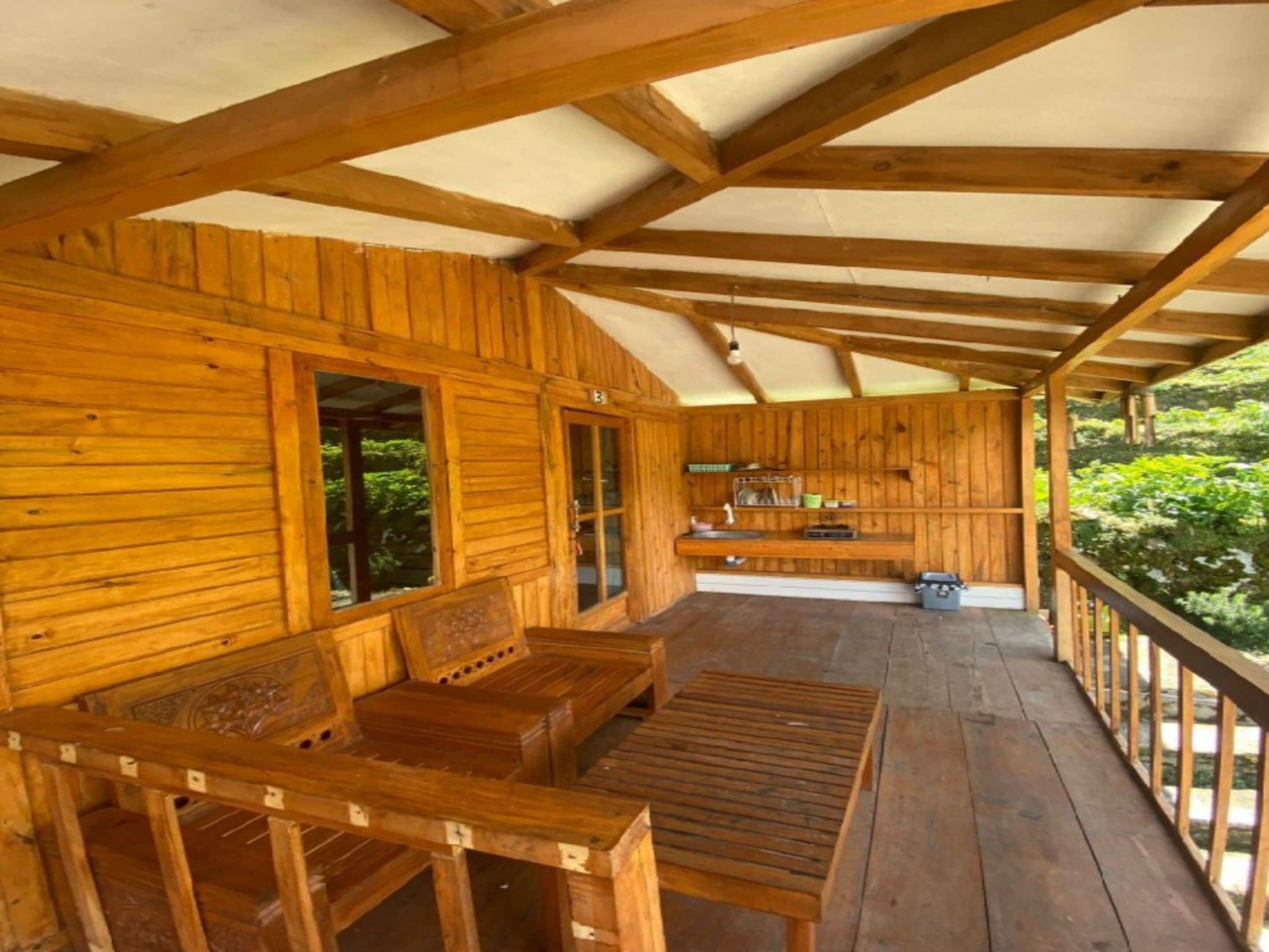 Una cabaña de madera con porche cubierto, bancos integrados y una pequeña cocina.