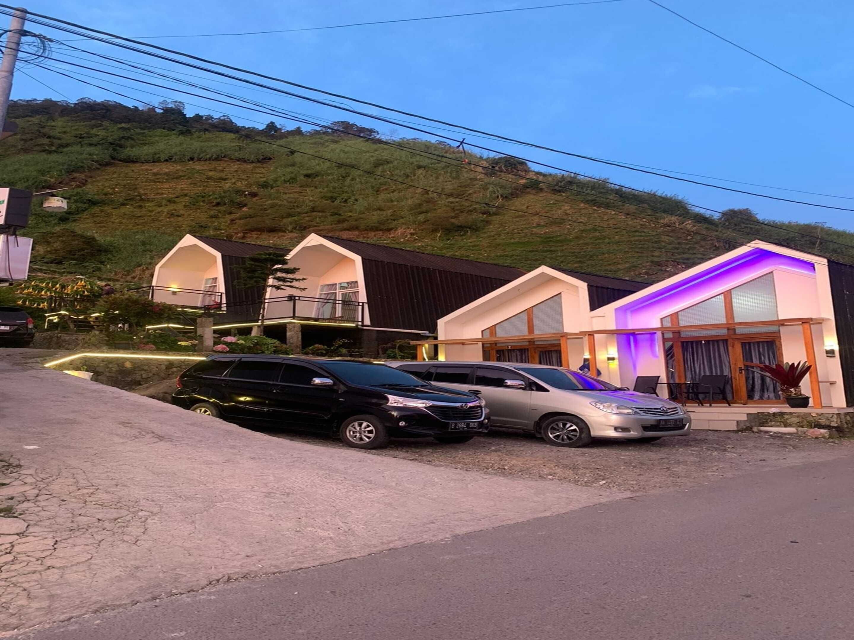 Um edifício moderno com telhado inclinado e varanda, localizado numa encosta com entrada pavimentada e carros estacionados.