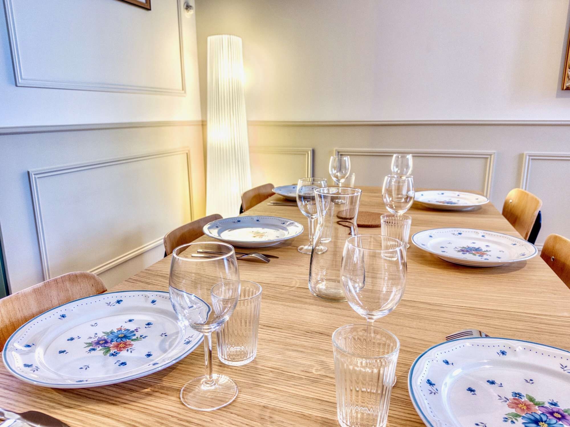 Ein Esstisch mit Geschirr mit Blumenmuster, klaren Trinkgläsern und Besteck, nebenan eine hohe, gerippte Lampe.
