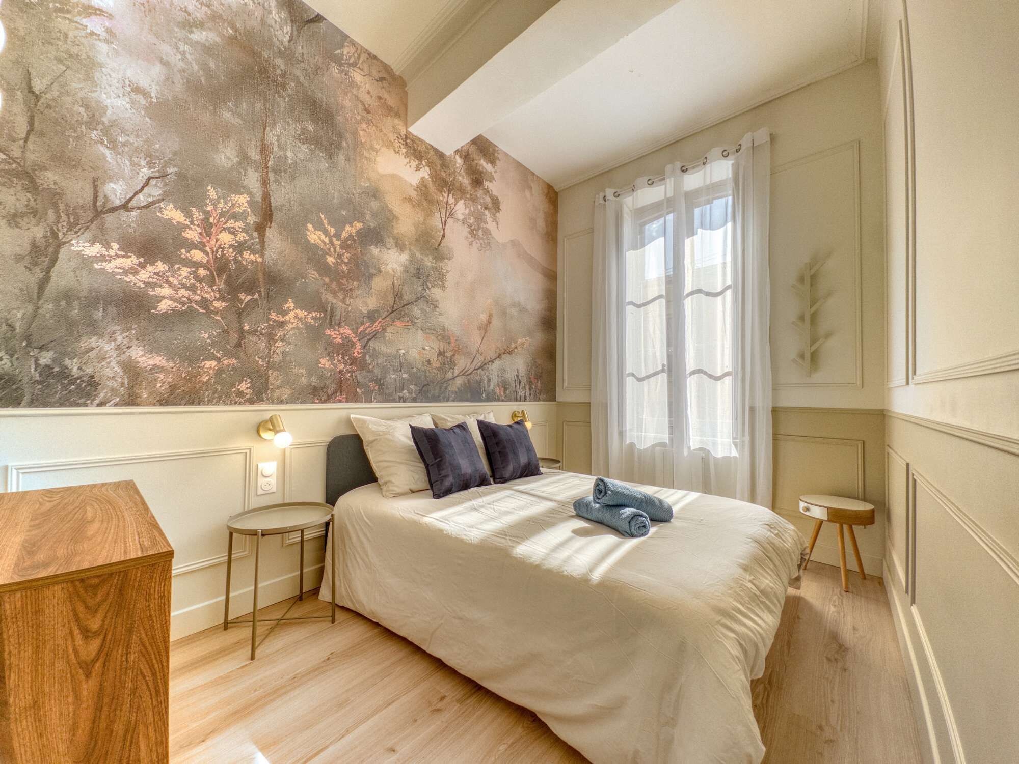 Ein Schlafzimmer mit einer großen Wandmalerei, einem Bett mit Kissen, einem Nachttisch, einer Holzkommode und einem Fenster mit Vorhängen.