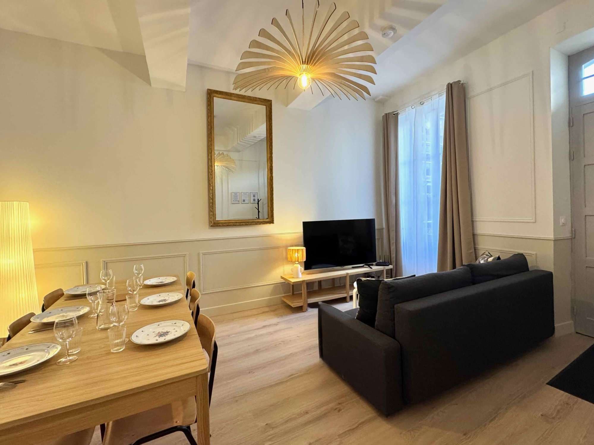 A modern living room with a wooden dining table set for a meal, a black sofa, a flat-screen TV, and a large mirror on the wall.