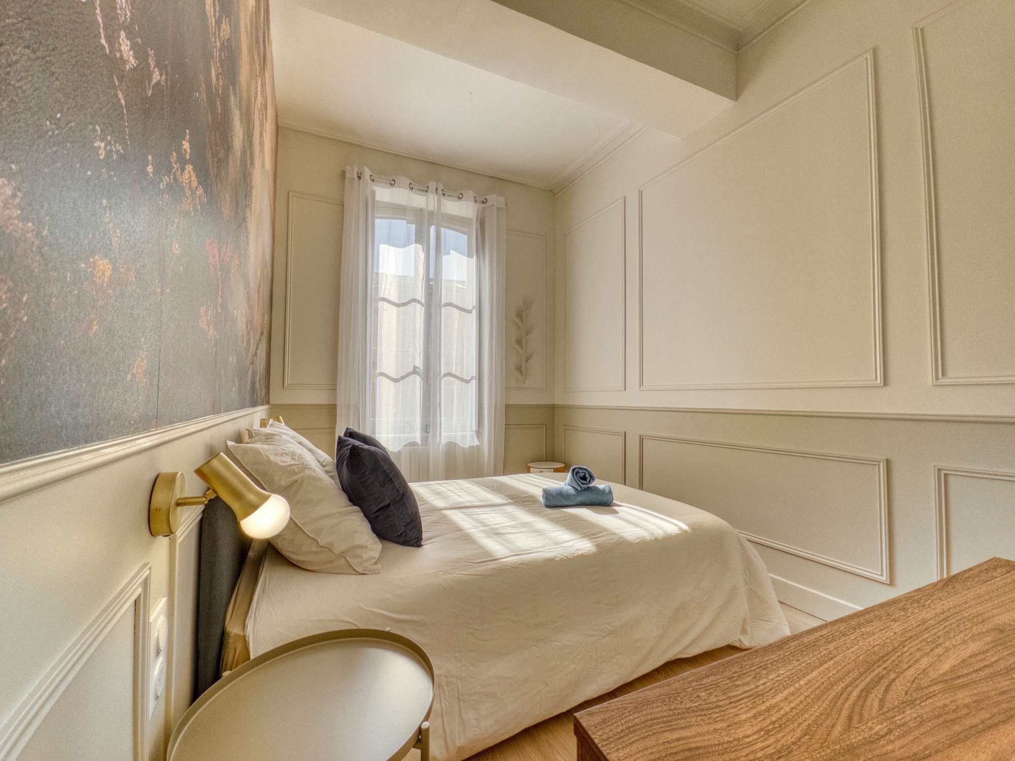A bedroom with a bed, a bedside lamp, a window with curtains, and a wooden headboard.