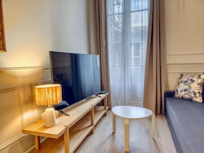 Un salon moderne avec un téléviseur à écran plat posé sur un meuble en bois, une petite table ronde, un canapé agrémenté d’un coussin à motifs et des rideaux légers qui laissent passer la lumière naturelle.