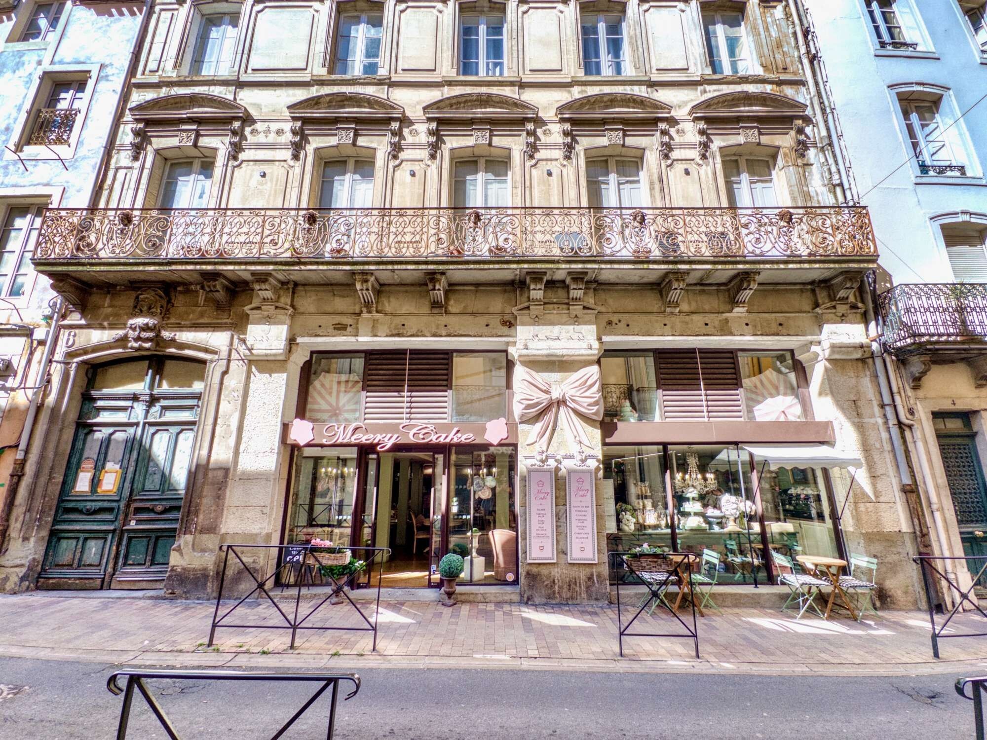 A building with a bakery named 'Meery Cake' on the ground floor, featuring a large window display and outdoor seating.