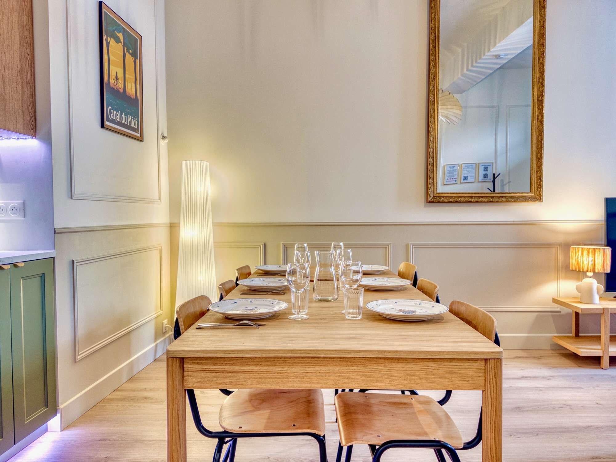 Un coin repas avec une table en bois dressée pour six personnes, entourée de chaises, agrémenté d’un grand lampadaire et d’un miroir au mur.