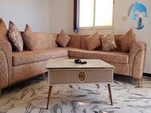 A living room with a beige sectional sofa, a marble coffee table, and a window with blinds. - Chott Mariem Apartment Sousse (Chott Mariem)