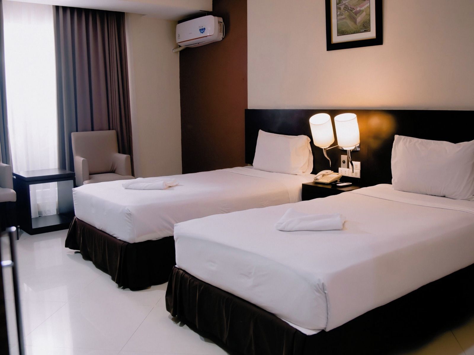 Quarto de hotel com duas camas, uma cadeira, mesa de cabeceira com abajur e ar condicionado na parede.