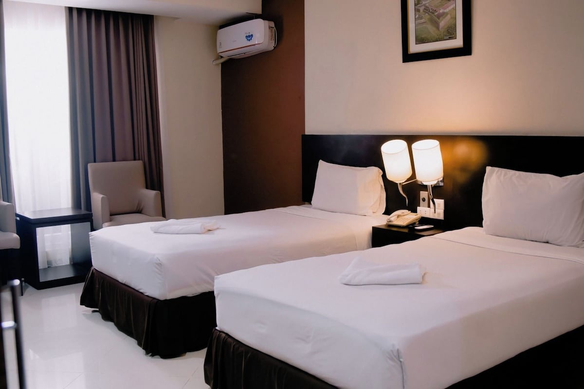 A hotel room with two beds, a chair, a bedside table with a lamp, and an air conditioning unit on the wall.
