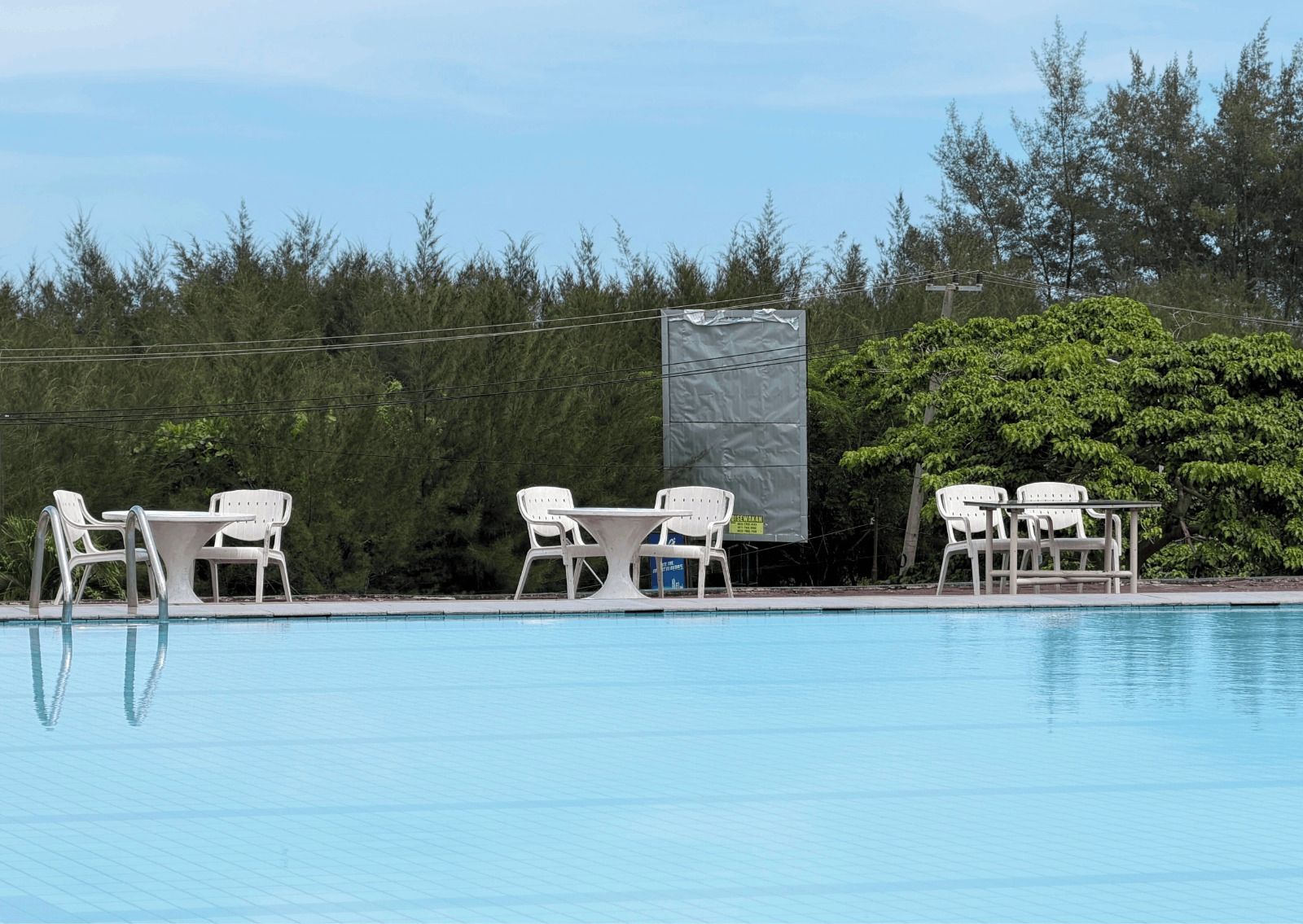 A swimming pool with white lounge chairs and a table, surrounded by trees and a covered area.