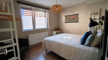 A bedroom with a bed, a bedside table, a hanging light fixture, a window with blinds, and a radiator.