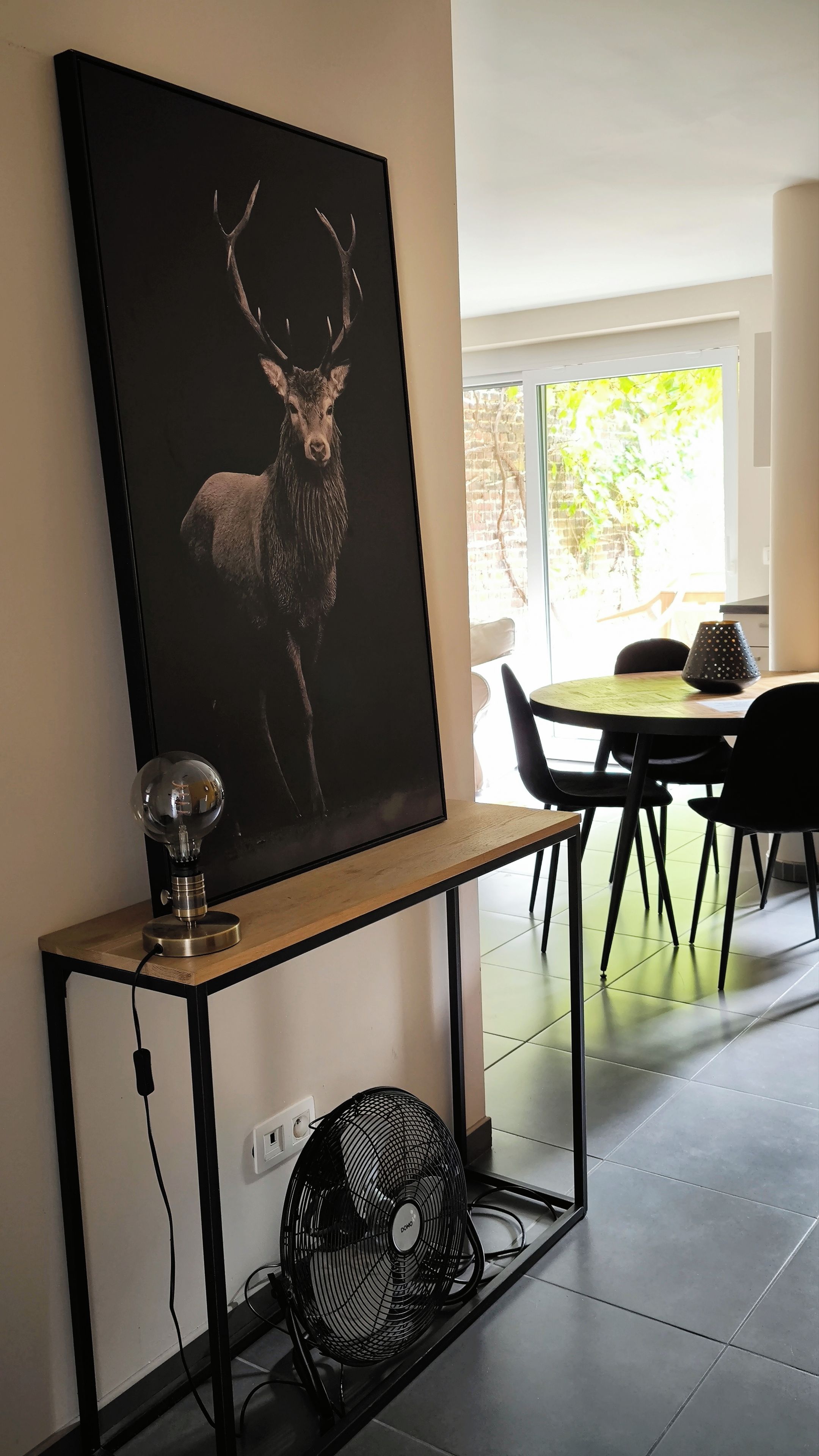 A dining area with a table and chairs, a wall-mounted picture of a deer, a lamp, and a fan on a console table.