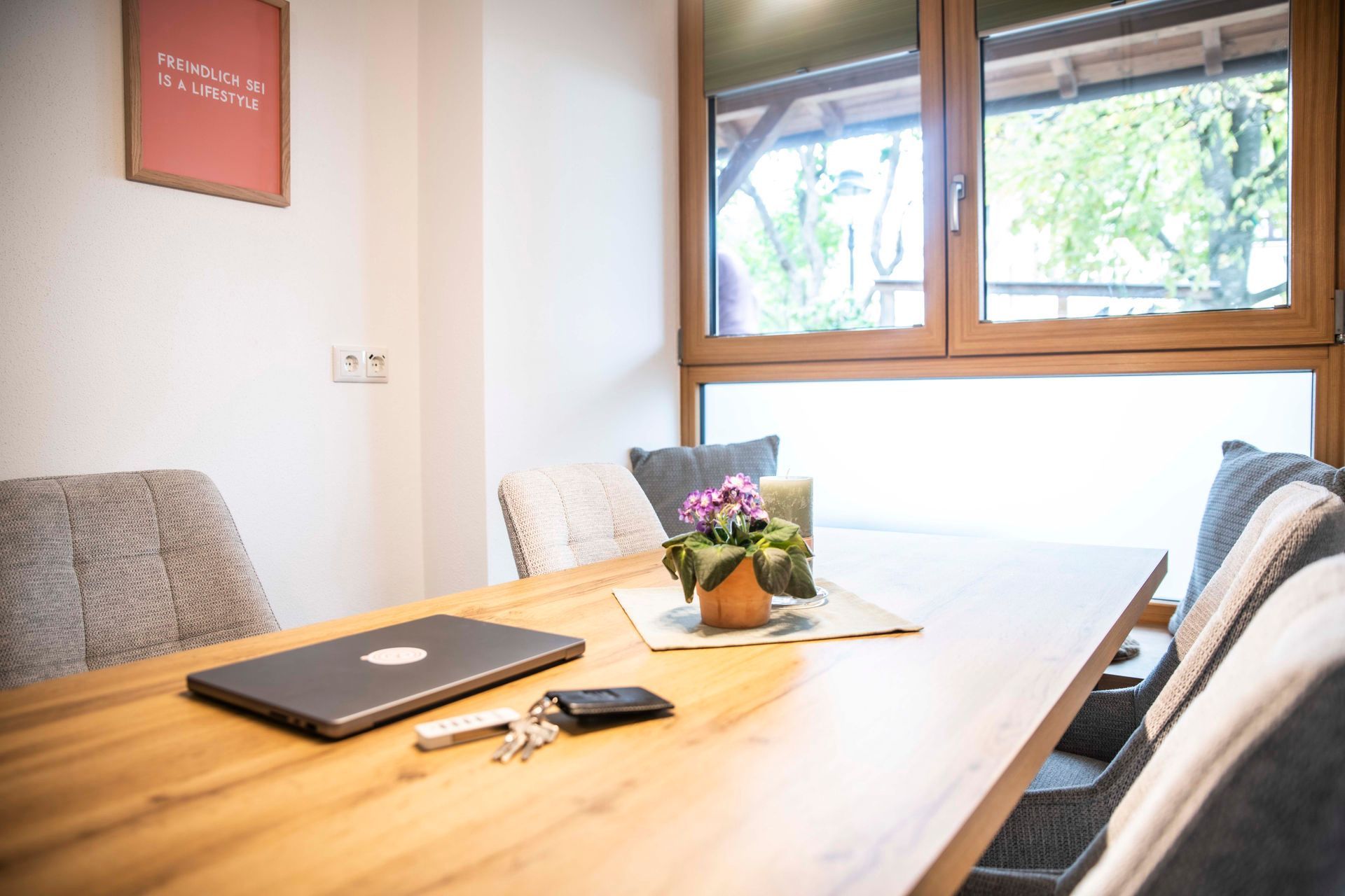 Ein Zimmer mit einem Holztisch, einem Laptop, einer Vase mit Blumen und einem Fenster mit Blick auf Grünflächen.