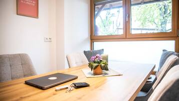 Ein Zimmer mit einem Holztisch, einem Laptop, einer Vase mit Blumen und einem Fenster mit Blick auf Grünflächen.