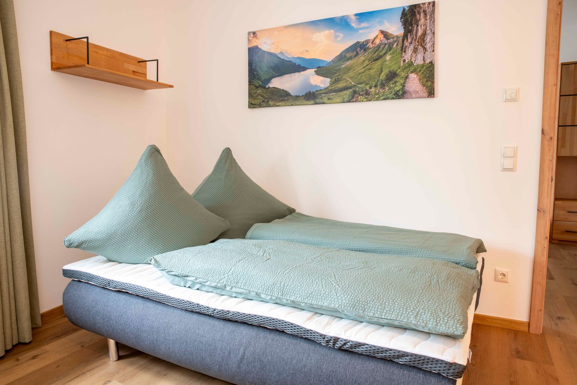 Ein Bett mit Kopfteil, ein an der Wand befestigtes Regal und ein Gemälde einer Berglandschaft.