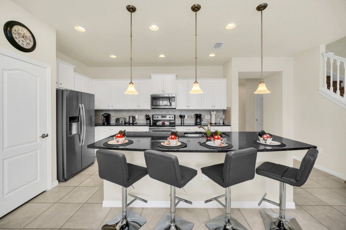 Una cocina moderna con una isla central, taburetes de bar, electrodomésticos de acero inoxidable, iluminación colgante y armarios blancos.