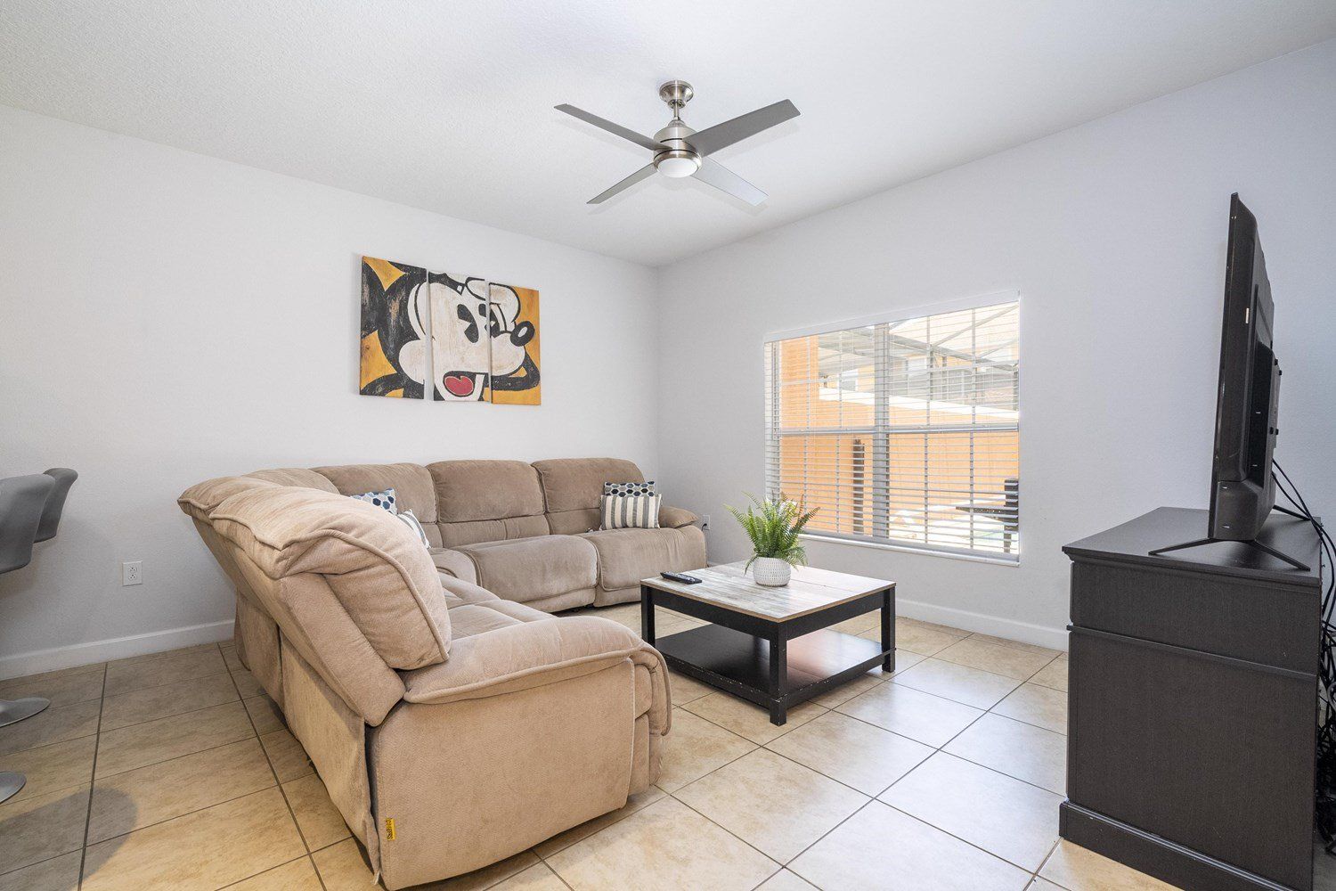 A living room with a beige sofa, a coffee table, a ceiling fan, a TV, and a window with blinds.