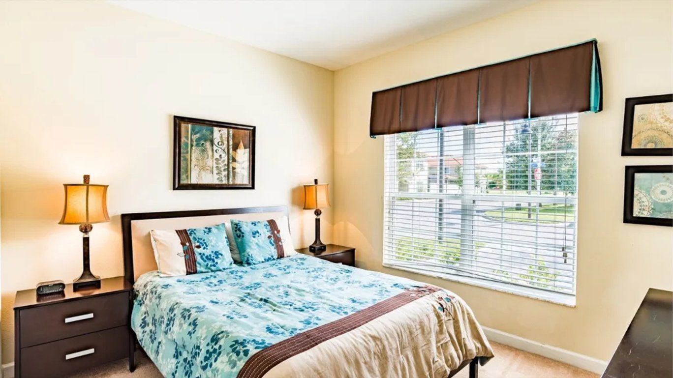 A bedroom with a bed, two bedside tables, a window with blinds, and artwork on the walls.