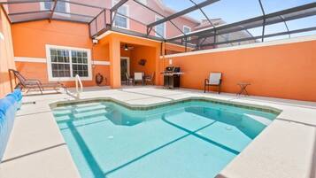 An indoor swimming pool with a glass ceiling, surrounded by orange walls and a patio area with chairs and a barbecue.