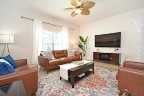 A living room with a brown leather sofa, a coffee table, a TV on the wall, and a large window with curtains. - Paradise Palms - Ocean 279 - 6530 (Kissimmee)