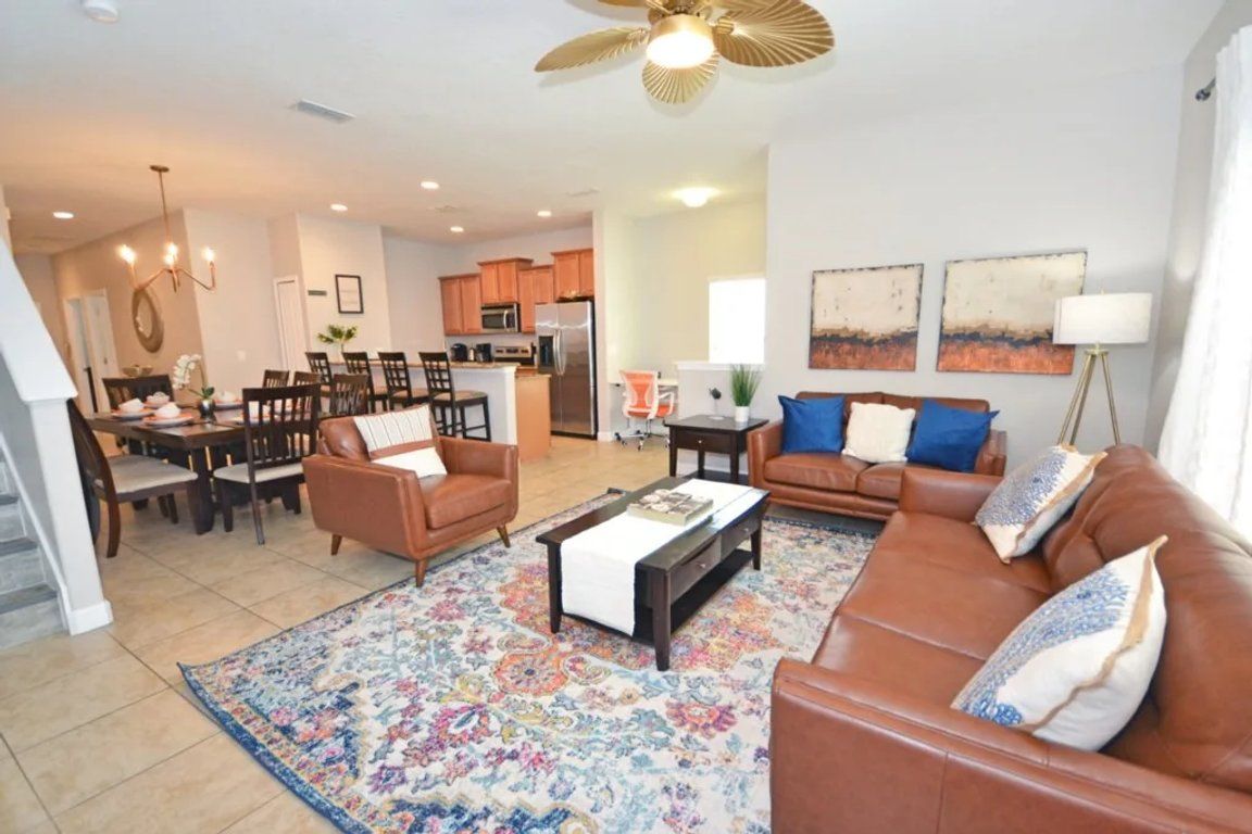 A spacious living room with a brown leather sofa, a coffee table, and a patterned rug. There is a dining area with a table and chairs, and a kitchen in the background.