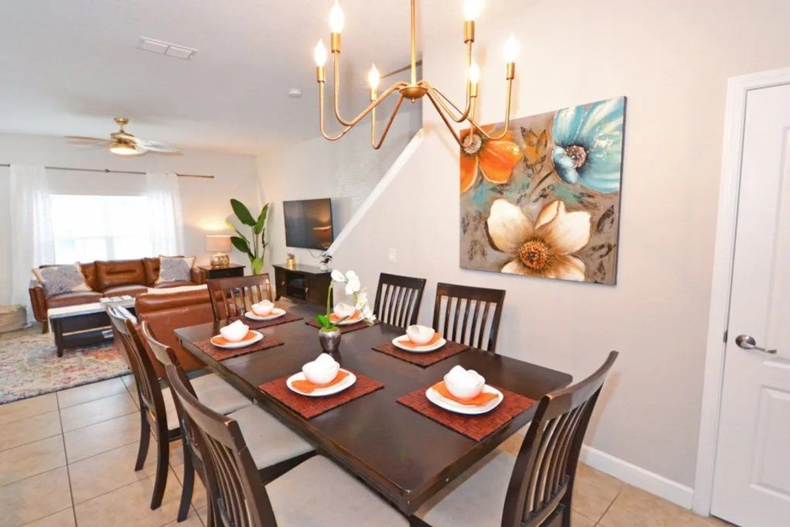 A dining area with a dark wooden table set for four, surrounded by chairs. A chandelier hangs above the table, and a large floral painting is on the wall.