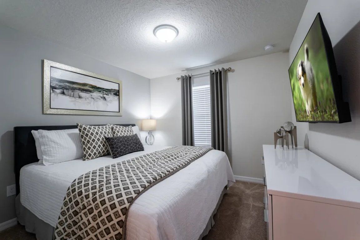 Un dormitorio con una cama, una mesita de noche, un televisor montado en la pared y un cuadro enmarcado sobre la cama.