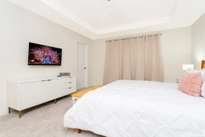 A bedroom with a bed, a television mounted on the wall, a bedside table, and a window with curtains. - Solara Resort - 6766 (Kissimmee)