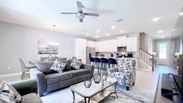A modern living room with a sofa, coffee table, and patterned armchair. There is a ceiling fan, recessed lighting, and a kitchen area in the background.