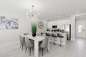 A modern kitchen with white cabinets, a stainless steel refrigerator, and a dining area with a white table and grey chairs. - Reunion - 7697 (Kissimmee)