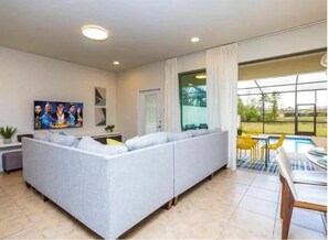 A modern living room with a sectional sofa, a flat-screen TV, and a dining area with yellow chairs. - Windsor at Westside - 11458 (Kissimmee)