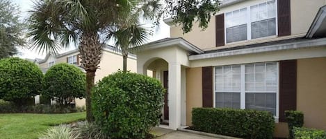 A residential building with a front entrance, a covered porch, and a well-maintained lawn with various shrubs and a palm tree.