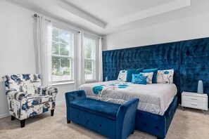 A bedroom with a large bed, a blue upholstered bench, a white nightstand, and a floral armchair. - Solara Resort - 7868 (Kissimmee)