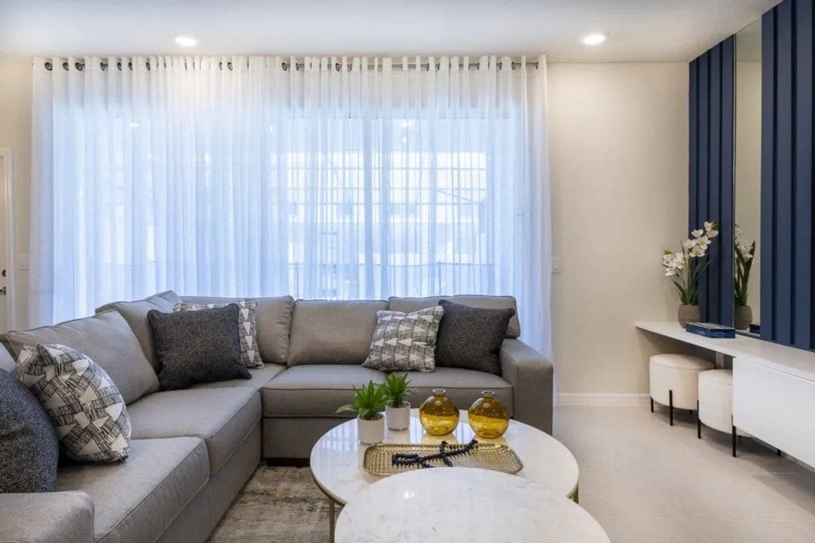 A modern living room with a sectional sofa, a round coffee table, and a large window with sheer curtains.