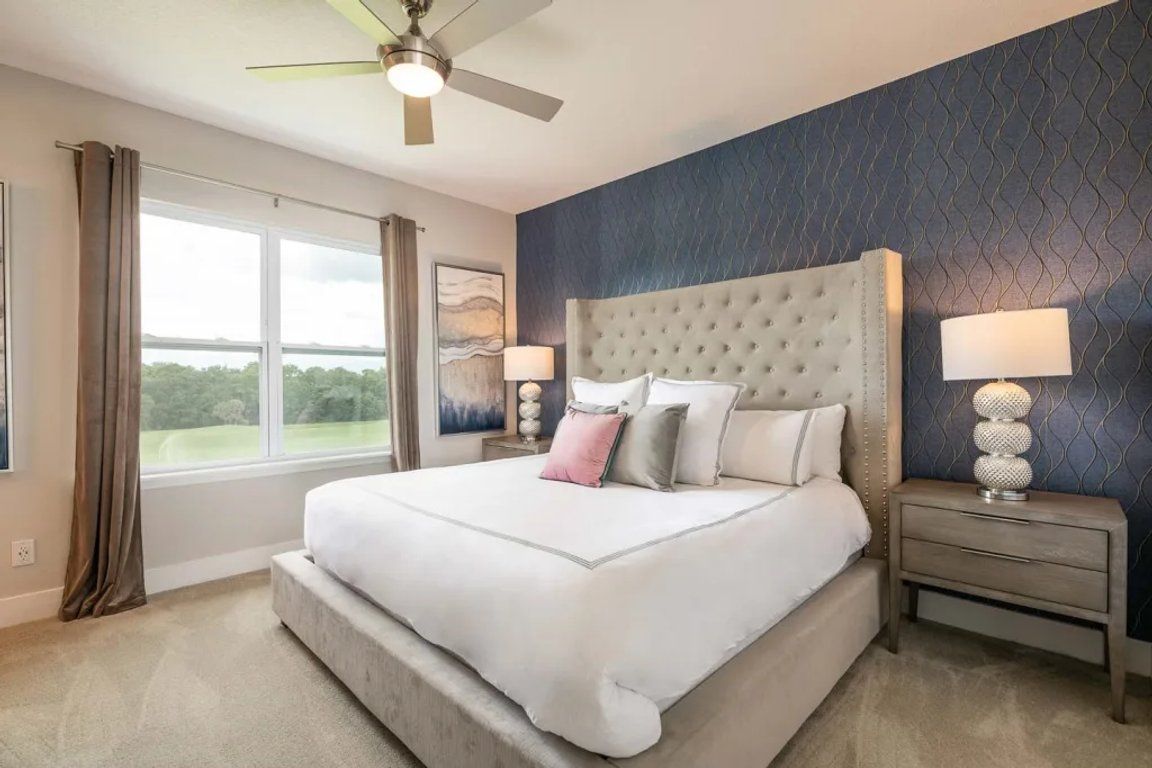 A bedroom with a large bed, a ceiling fan, a nightstand with a lamp, and a view of the outdoors through a window.