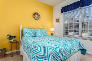 A bedroom with a bed, a nightstand, a lamp, a window with blinds, a wall with a wreath, and a small table with a plant. - Windsor Palms - 11478 (Kissimmee)