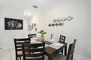 Un comedor con una mesa dispuesta para cuatro personas, rodeada de sillas, un jarrón con una planta y un adorno en la pared.