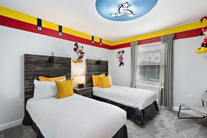 Habitación de hotel con dos camas, cabecera de madera, un adhesivo de pared con Mickey Mouse y una ventana con cortinas.