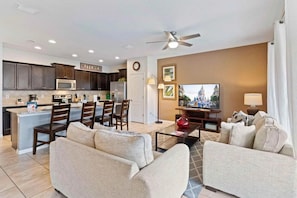 A modern living room with a sofa set, a dining area with chairs, a kitchen with cabinets and appliances, and a television on the wall. - Windsor at Westside - 25962 (Kissimmee)