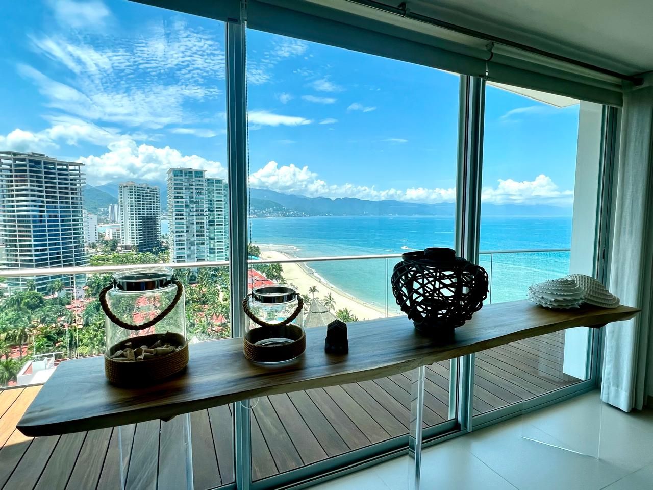 Een balkon met uitzicht op een strand en stadsbeeld, met een houten tafel en decoratieve objecten.