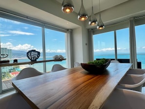 A dining area with a wooden table, white chairs, and a view of the ocean. - Luxury Oceanfront Corner Condo | Icon Vallarta (Puerto Vallarta)